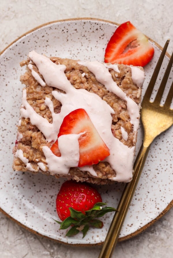 A square piece of oatmeal bake topped with white icing and sliced strawberries on a speckled plate, with a gold fork beside it.
