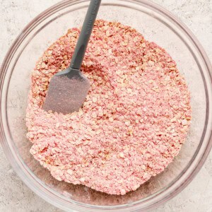A glass bowl filled with pink-tinted oats and a gray spatula resting inside, placed on a light textured surface.
