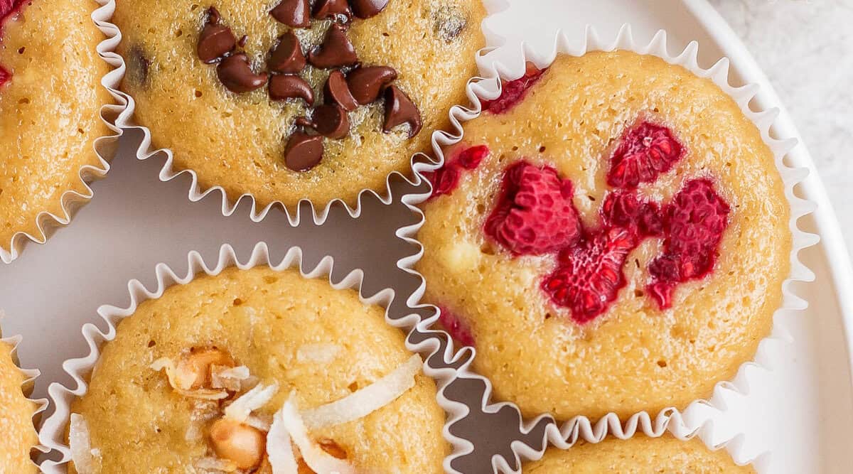 Three muffins on a white plate, each topped with chocolate chips, chopped raspberries, and coconut shreds—a perfect treat for The Cream Blog enthusiasts.