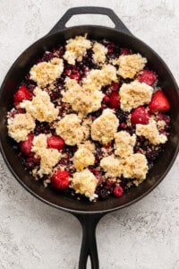 A cast iron skillet filled with mixed berries topped with dollops of crumbly dough and brown sugar, ready for baking.