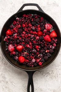 A cast iron skillet filled with a mix of frozen berries, including strawberries, raspberries, blackberries, and blueberries, on a light textured surface.