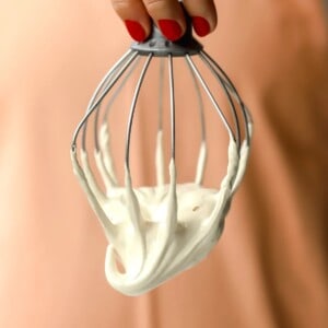 A hand with red nail polish holds a metal whisk covered in thick, maple marshmallow fluff against a peach-colored background.