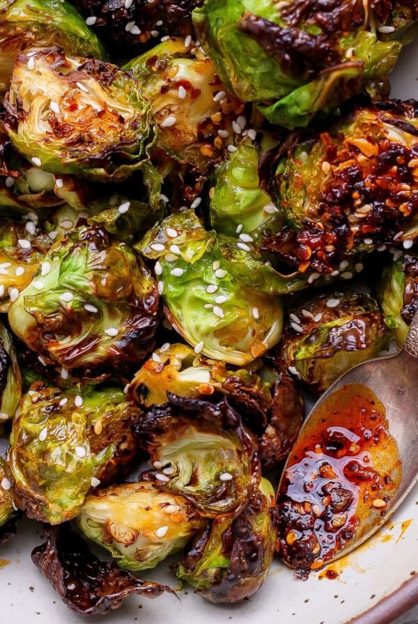 Roasted Brussels sprouts with a glossy chili sauce, garnished with sesame seeds, served in a bowl with a spoon.