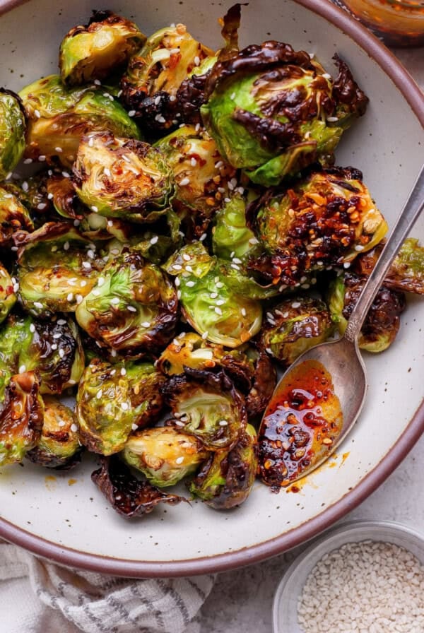 A bowl of roasted Brussels sprouts garnished with sesame seeds, served with a spoon and chili oil.