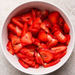 A white bowl filled with sliced fresh strawberries sits on a light textured surface.