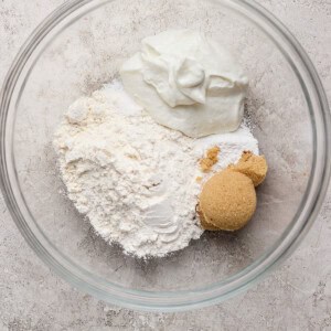 A glass bowl containing flour, brown sugar, and a scoop of white yogurt on a light textured surface.