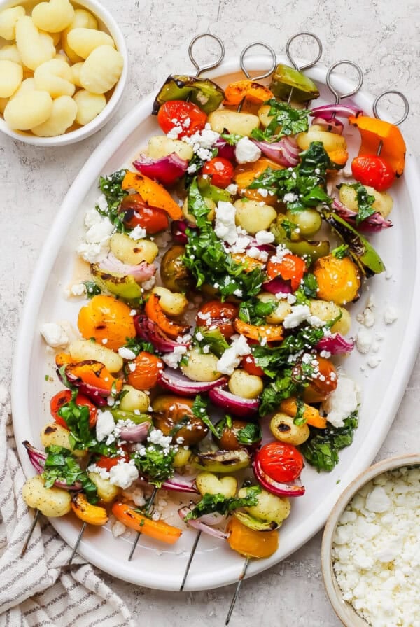 A white platter with vegetable and gnocchi skewers, topped with chopped herbs and crumbled cheese, is pictured next to small bowls of cooked gnocchi and crumbled cheese.