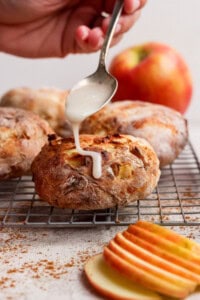 A hand drizzles white icing from a spoon onto a round baked pastry with apple pieces, resting on a cooling rack. Sliced apples and a whole apple are in the background.