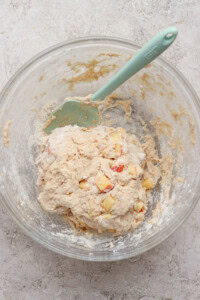 A glass bowl containing dough mixed with small apple chunks and a green spatula on a light textured surface.