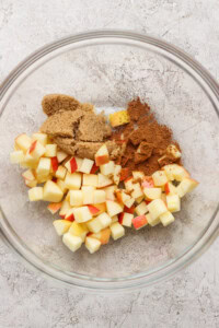 A glass bowl containing diced apples, brown sugar, and ground cinnamon on a light textured surface.