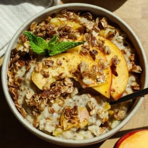 A bowl of oatmeal topped with granola, peach slices, and a sprig of mint, with a spoon placed inside.