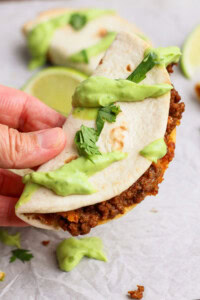A hand holds a taco filled with ground beef and topped with green sauce and chopped cilantro on a light surface.