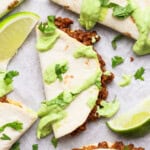 Taco halves with ground meat filling, topped with green sauce and cilantro, served with lime wedges on a white surface.