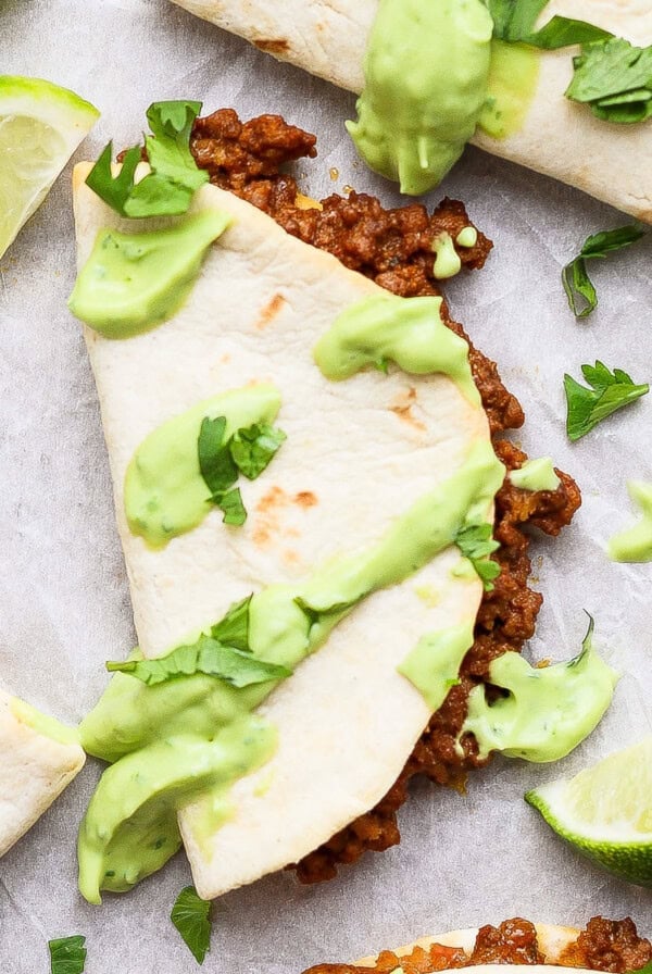 Taco halves with ground meat filling, topped with green sauce and cilantro, served with lime wedges on a white surface.