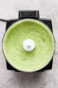 Top view of a food processor containing smooth, creamy green sauce or dip, likely a blended avocado or herb mixture.
