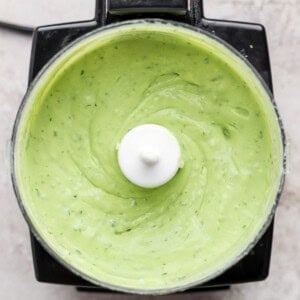 Top view of a food processor containing smooth, creamy green sauce or dip, likely a blended avocado or herb mixture.