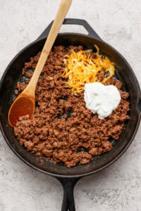 Ground beef in a cast iron skillet with shredded cheese and a dollop of sour cream, being stirred with a wooden spoon.