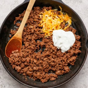 Ground beef in a cast iron skillet with shredded cheese and a dollop of sour cream, being stirred with a wooden spoon.