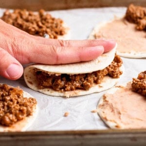 A hand folds a tortilla filled with ground meat mixture on a baking sheet lined with parchment paper, with more tortillas being assembled nearby.