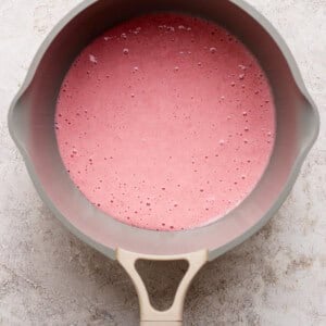 A beige saucepan filled with a pink liquid mixture sits on a light textured surface, viewed from above.