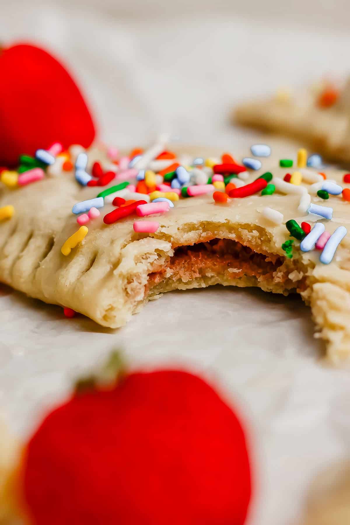 A close-up of a frosted, sprinkle-topped pastry with a bite taken out, showing a strawberry filling. Whole strawberries are also visible in the background.