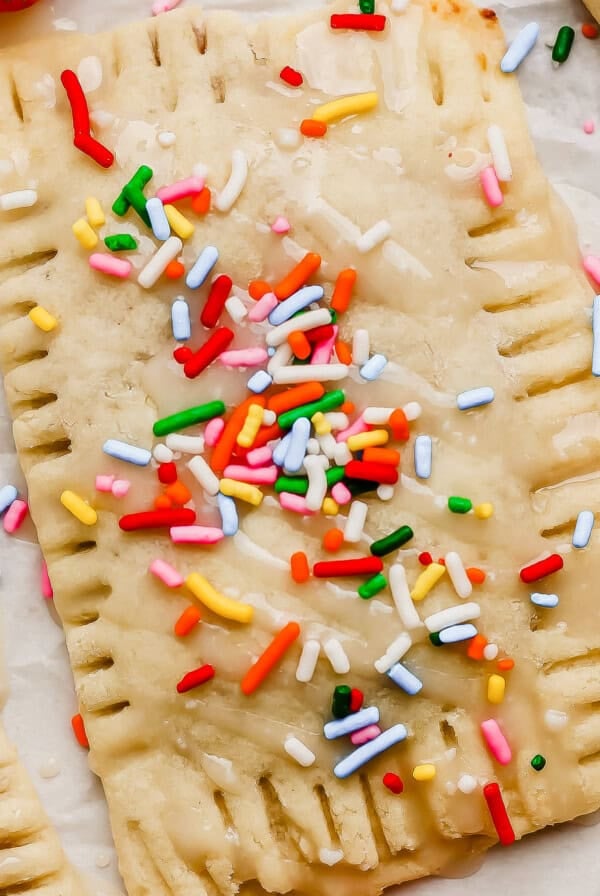 A rectangular pastry topped with icing and colorful sprinkles sits on parchment paper next to a fresh strawberry.