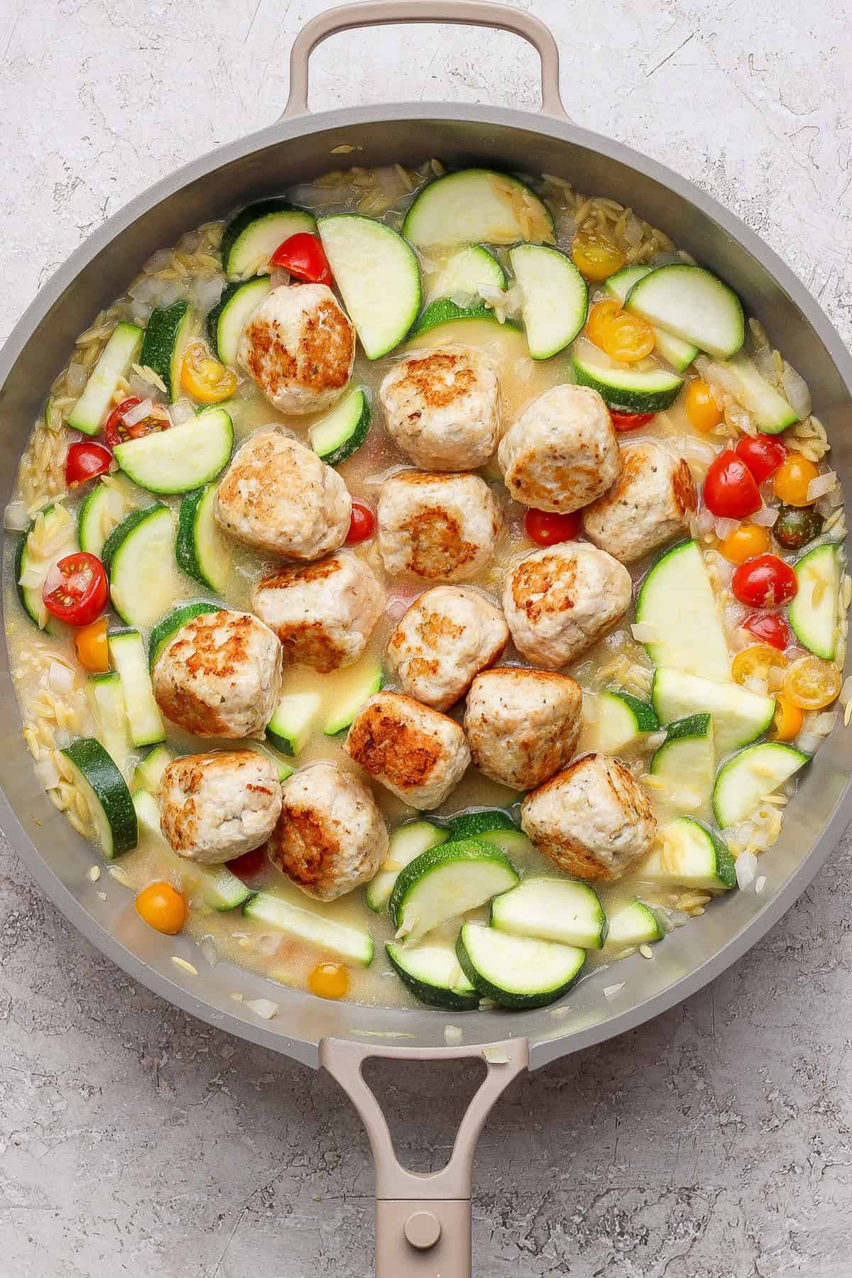 A skillet with browned meatballs, sliced zucchini, yellow squash, cherry tomatoes, and orzo in a light sauce on a gray surface.