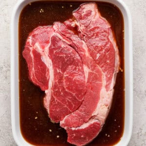 A raw beef steak sits in a white ceramic dish filled with brown marinade on a light textured surface.