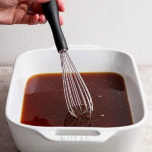 A hand uses a whisk to mix a dark liquid marinade in a white rectangular baking dish on a light surface.