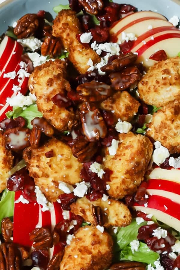 A salad with mixed greens, sliced red apples, breaded chicken pieces, pecans, dried cranberries, crumbled feta cheese, and a light drizzle of dressing.
