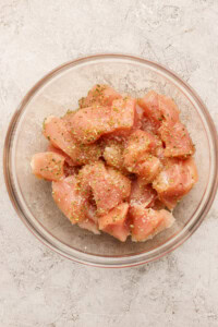 A glass bowl filled with raw, diced chicken breast pieces, seasoned with salt, pepper, and dried herbs, on a light textured surface.