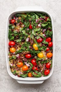 A white baking dish filled with fresh spinach, cherry tomatoes, chopped red onion, sun-dried tomatoes, and olives on a light textured surface.