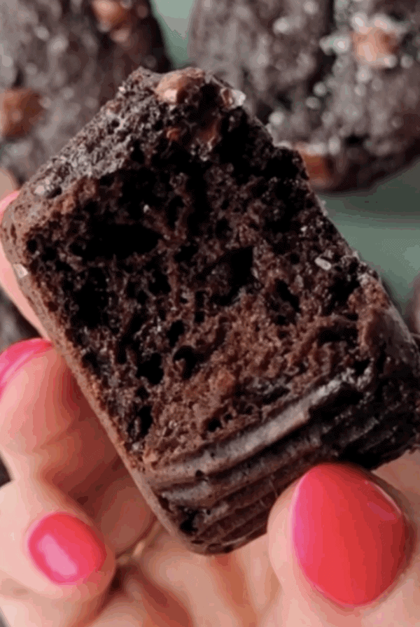 A hand with pink nail polish holds a bitten Kodiak protein muffin, revealing its moist, dense interior; more chocolate muffins are blurred in the background.