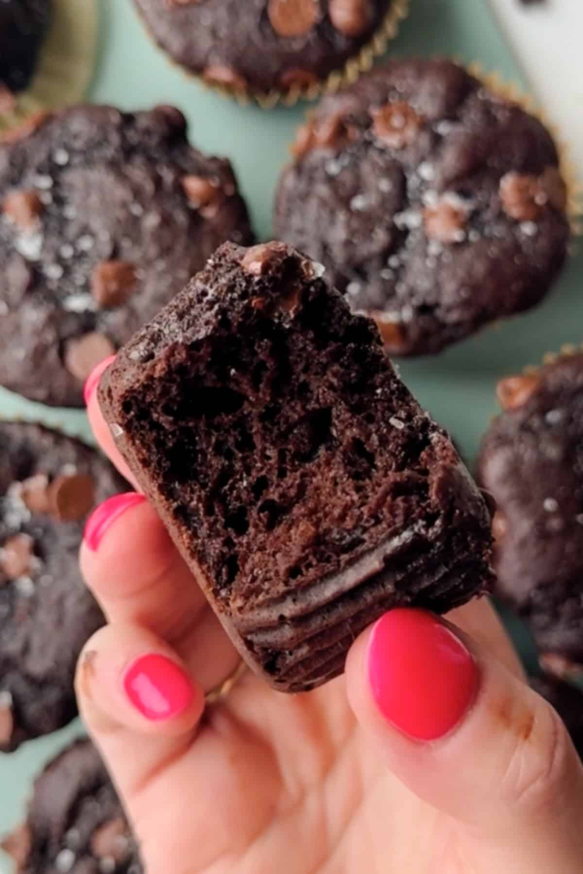 A hand with pink nail polish holds a partially eaten chocolate muffin, with more chocolate muffins visible in the background.