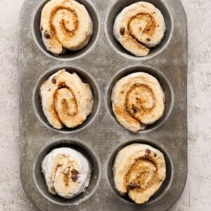 Six unbaked cinnamon rolls with chocolate chips arranged in a metal muffin tin on a textured surface.