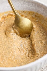 A gold spoon stirs a thick, light brown chia pudding mixture in a white bowl.