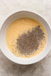 A white bowl containing whisked eggs with a pile of chia seeds on top, set on a light-colored textured surface.