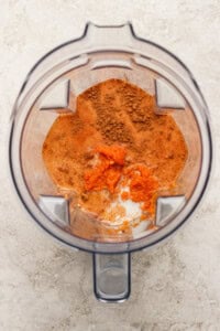 Overhead view of a blender containing orange puree, milk, and brown spices on a light-colored surface.