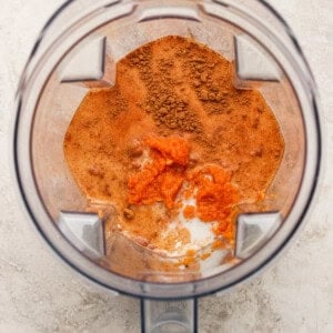 Overhead view of a blender containing orange puree, milk, and brown spices on a light-colored surface.