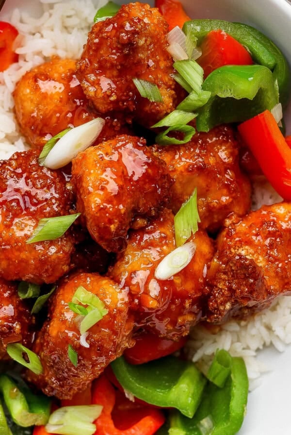 A bowl of white rice topped with crispy glazed chicken pieces, sliced green onions, and red and green bell peppers.