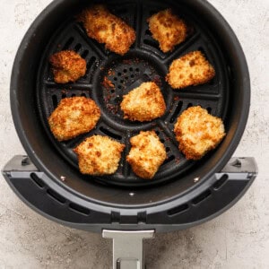 Eight breaded chicken nuggets are arranged in the basket of an air fryer, cooked to a golden brown color on a textured light surface.