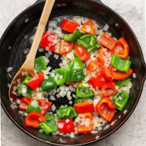 A cast-iron skillet with chopped red and green bell peppers and diced onions being sautéed, stirred with a wooden spoon.