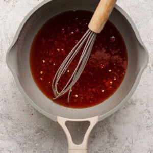 A saucepan containing a reddish-brown sauce with chili flakes, being whisked with a metal whisk on a textured light surface.
