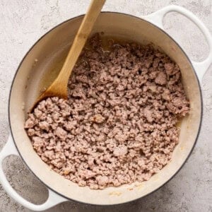 Cooked ground meat in a white pot with a wooden spoon, photographed from above on a light textured surface.