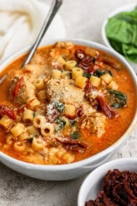 A bowl of soup with meatballs, ditalini pasta, spinach, sun-dried tomatoes, and grated cheese, served with a spoon.
