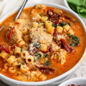 A bowl of soup with meatballs, ditalini pasta, spinach, sun-dried tomatoes, and grated cheese, served with a spoon.