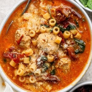 A bowl of tomato-based soup with meatballs, ditalini pasta, sun-dried tomatoes, spinach, and grated cheese on top.