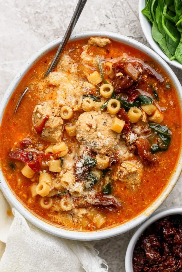 A bowl of tomato-based soup with pasta, chicken, spinach, sun-dried tomatoes, and grated cheese, with a spoon resting inside and fresh spinach on the side.