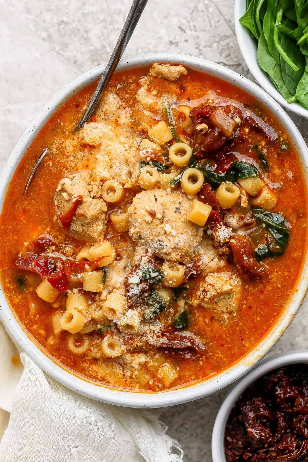 A bowl of tomato-based soup with pasta, chicken, spinach, sun-dried tomatoes, and grated cheese, with a spoon resting inside and fresh spinach on the side.