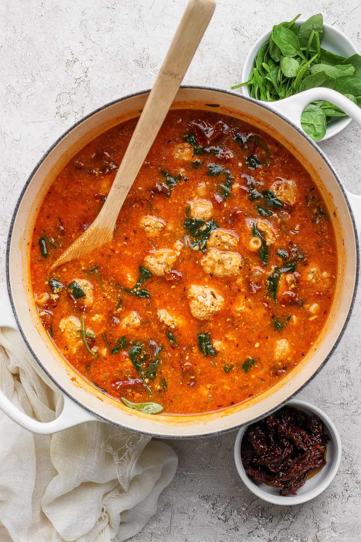 A pot of tomato-based soup with meatballs and spinach, a wooden spoon resting inside. Nearby are bowls of fresh spinach and sun-dried tomatoes.